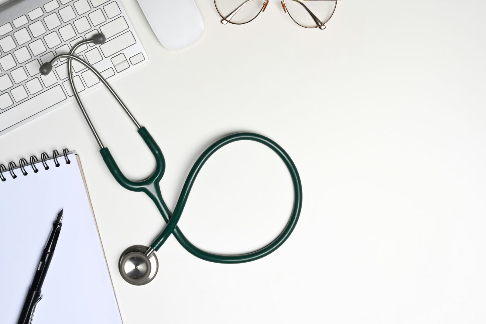 Stethoscope beside a blank notebook, fountain pen and glasses on a white desk