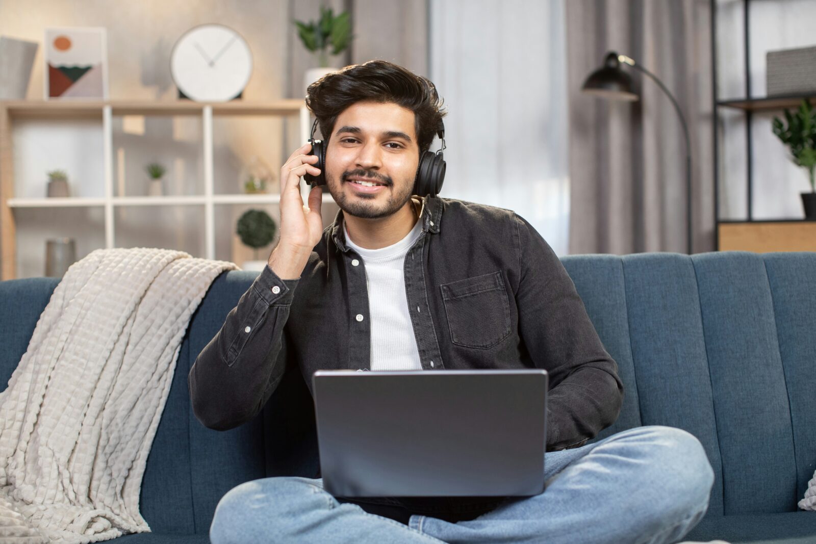 Young man wearing headphones and using a laptop on a blue sofa at home