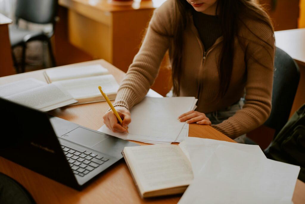 Learner writing notes at a desk beside open books and a laptop