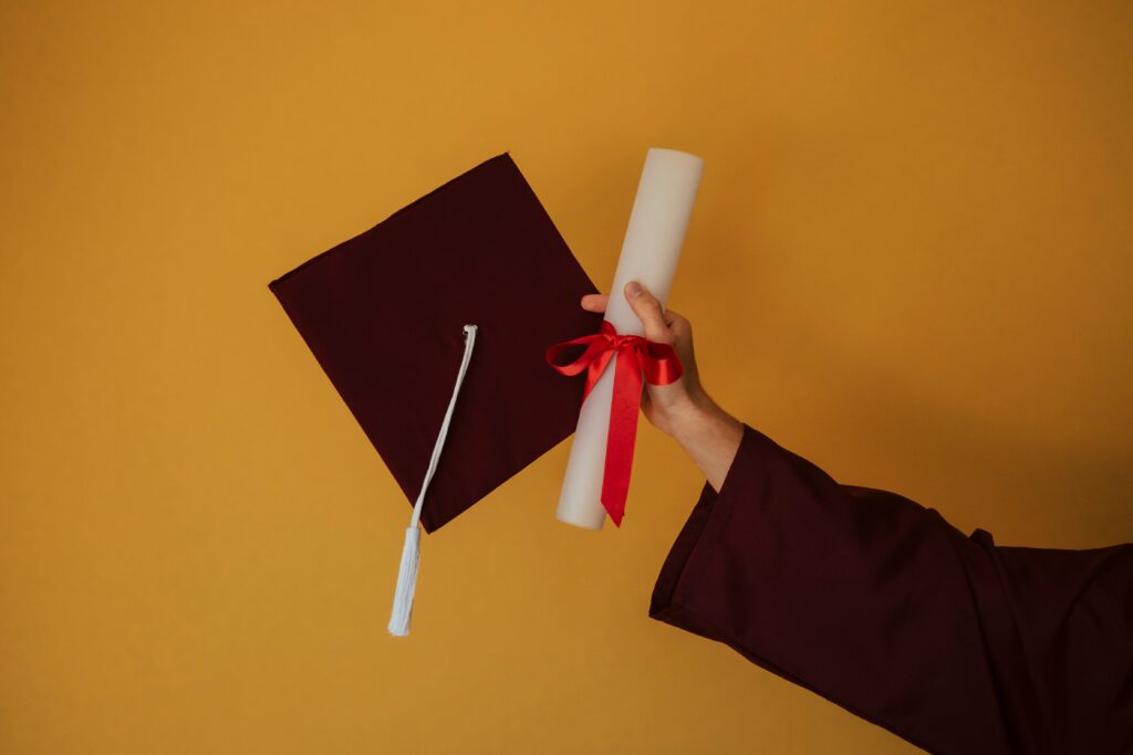 Hand holding a rolled degree certificate tied with red ribbon beside a graduation cap