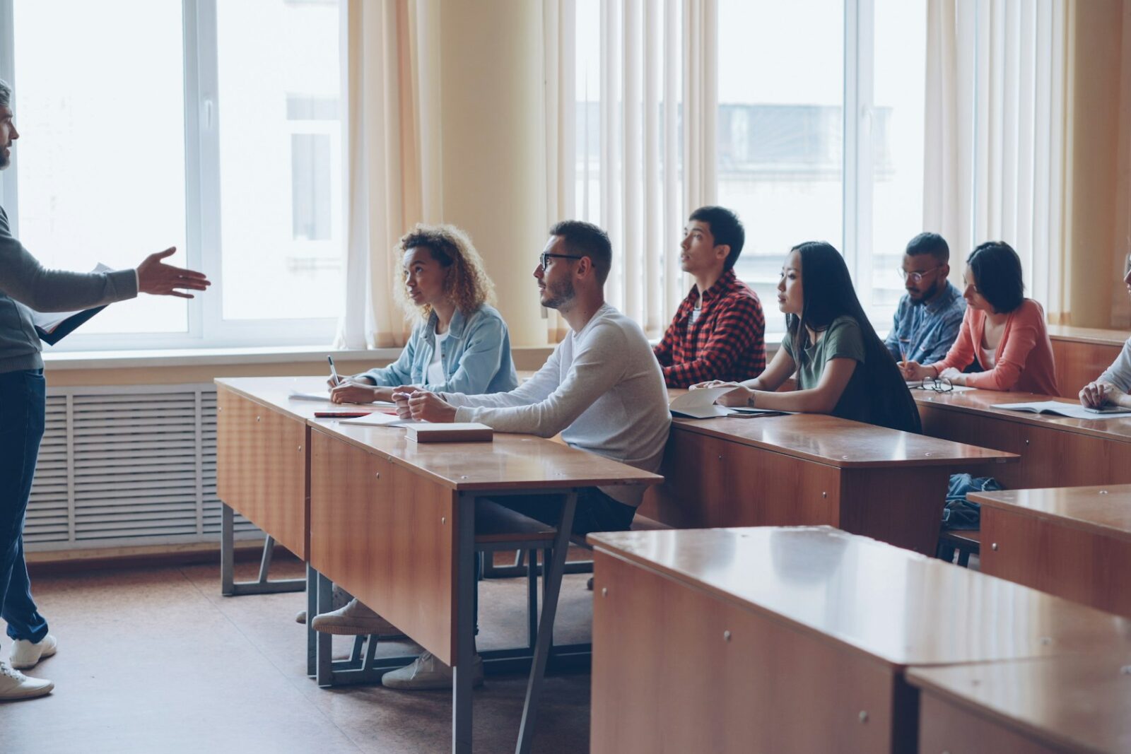 A teacher talking to a group of adult learners taking notes at tables in a bright classroom