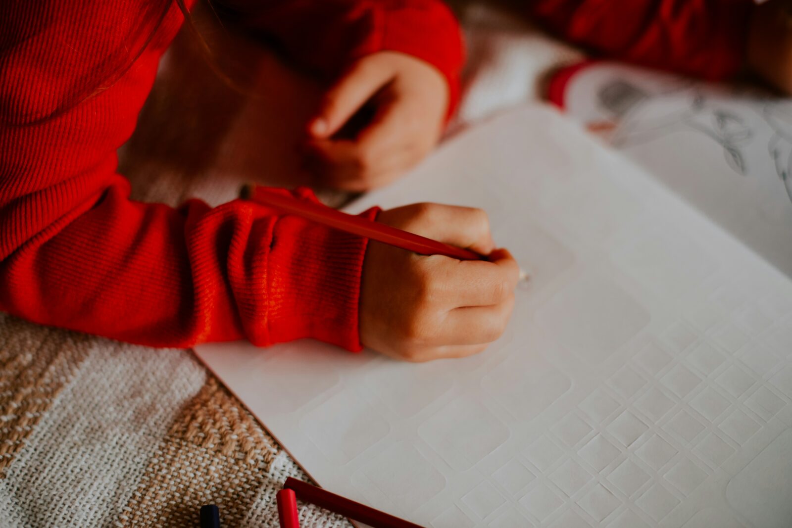 Child in a red jumper drawing with a red pencil on a sheet of white paper at home
