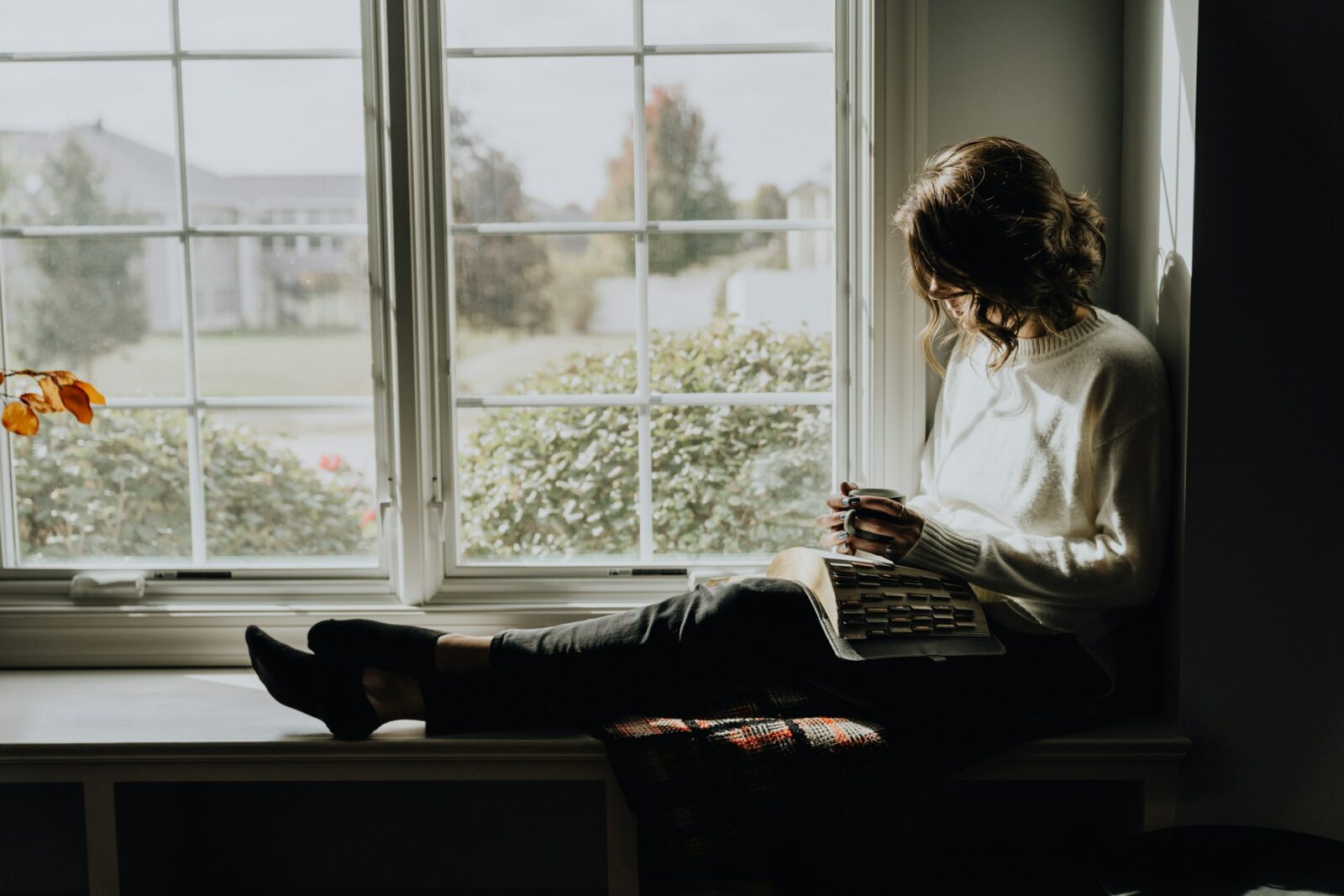 5 Books For Self-Help And Motivation A woman sitting on a window sill reading a book in soft daylight