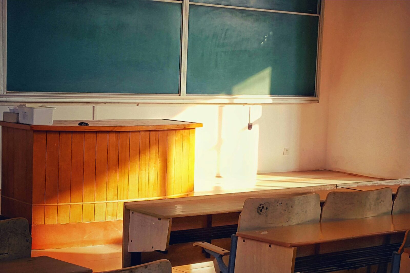 A Glimpse Of Government Schools In India Sunlit empty classroom with a wooden teacher's desk, worn student benches and green chalkboards