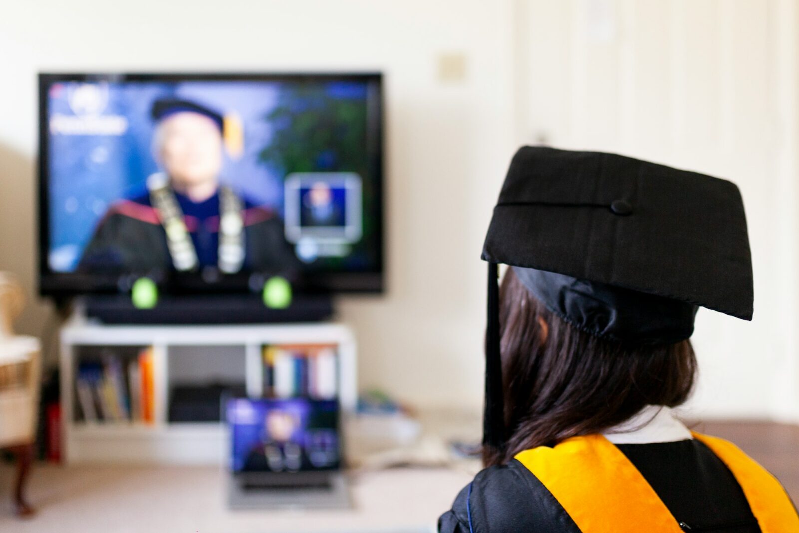 Graduate in cap and gown seen from behind watching an online graduation ceremony on a television