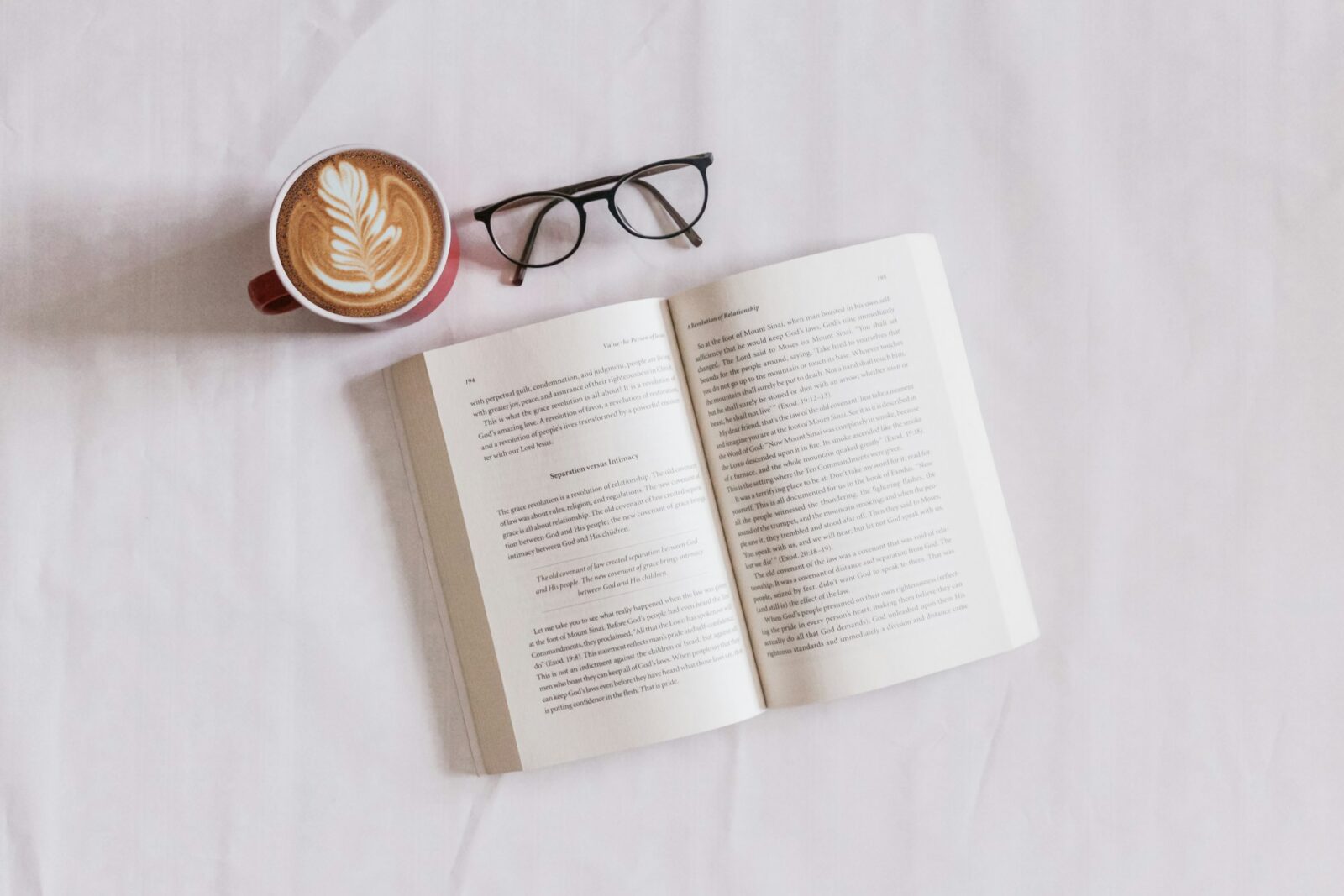 An open book beside a pair of glasses and a cup of cappuccino