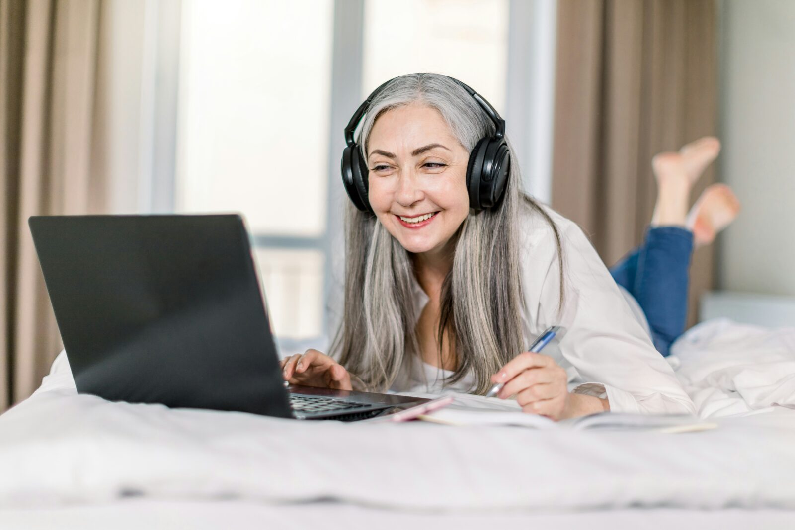 Top 5 Best Youtube channels to learn English Online Woman with grey hair wearing headphones takes notes beside a laptop on a bed