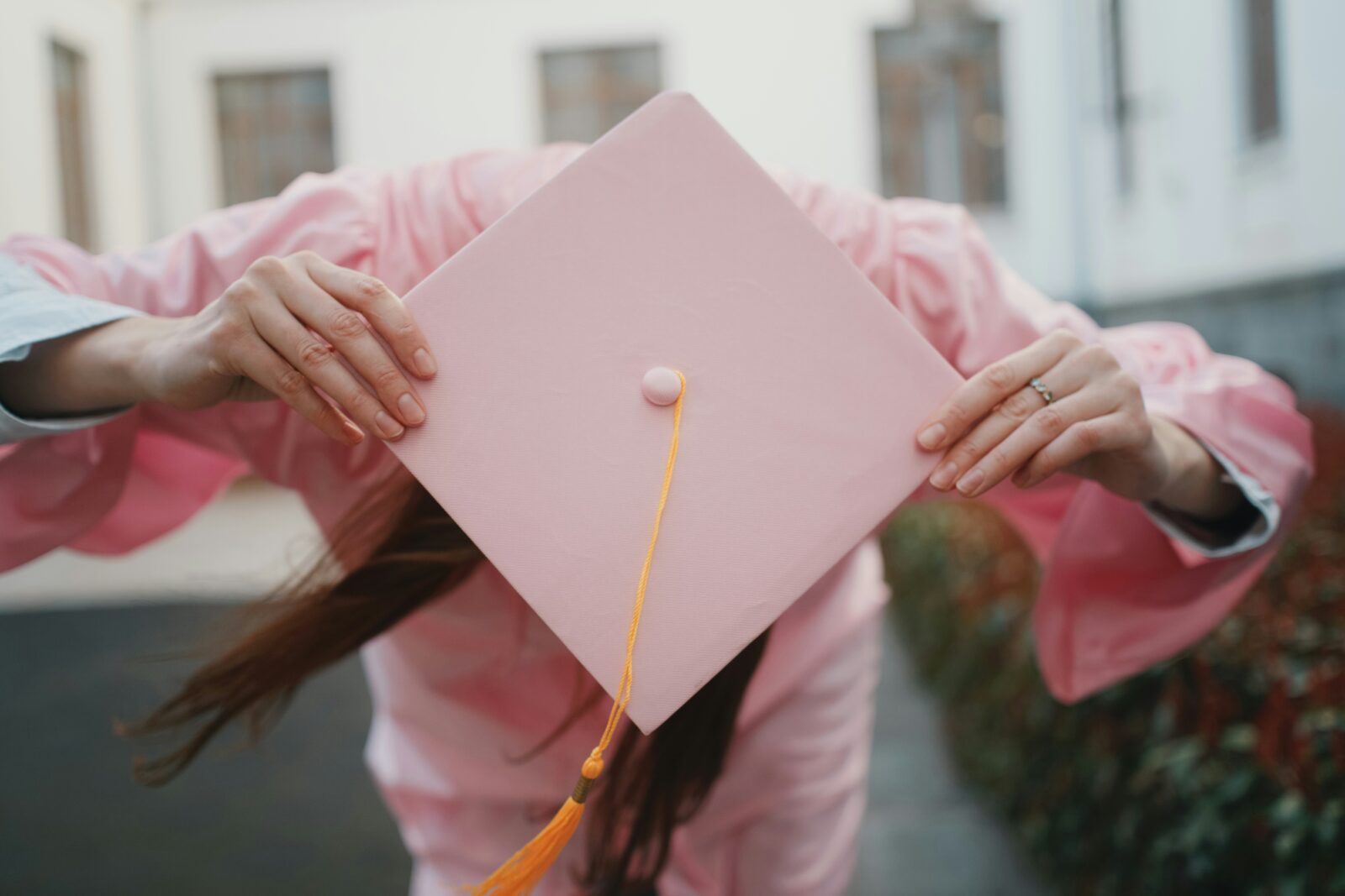 A graduate in a pink gown holds a pink mortarboard in front of her face